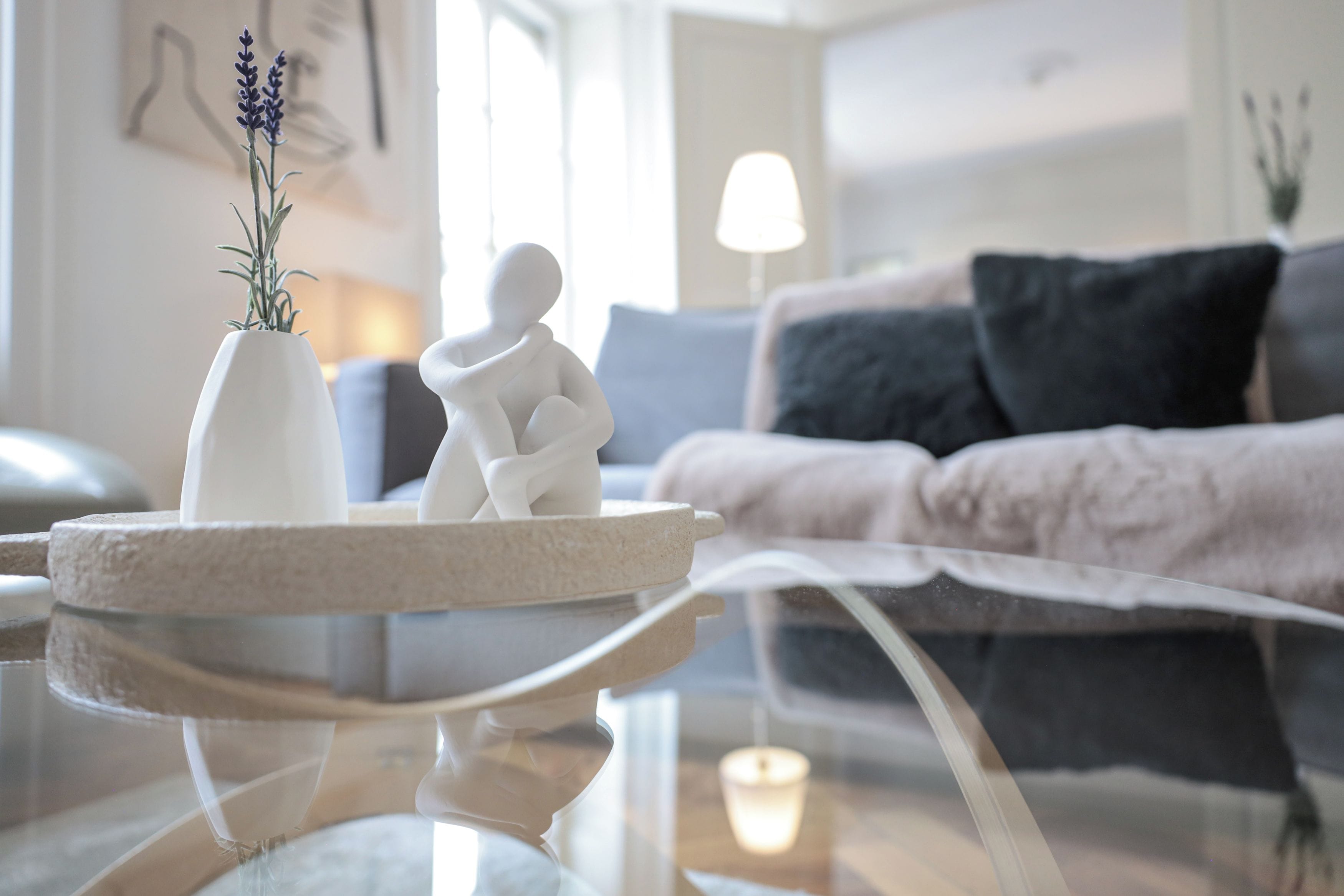 A modern living room with a glass coffee table, a white vase with a plant, a white sculpture, and a grey sofa with black cushions.