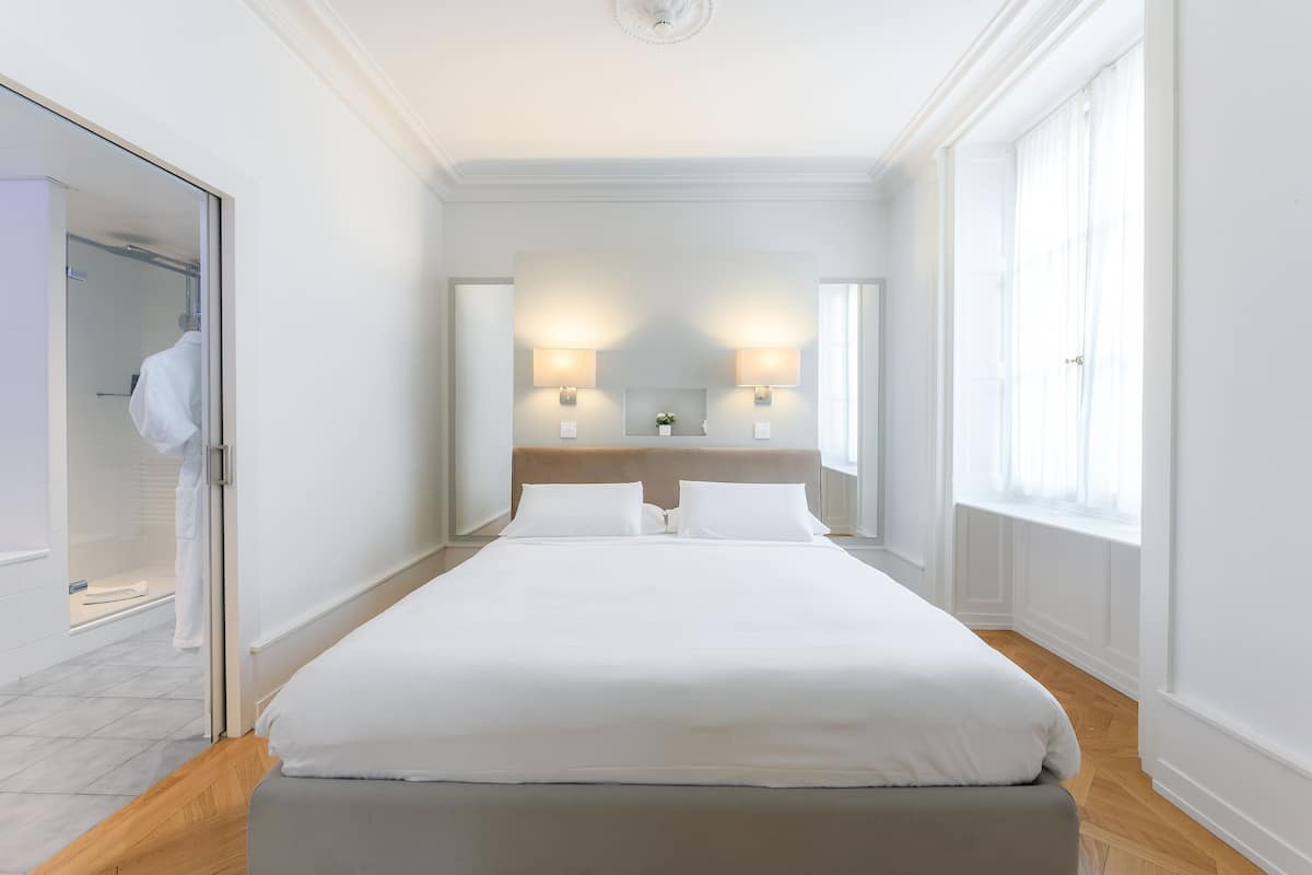 A hotel room with a bed, bedside lamps, a mirror, and a window.