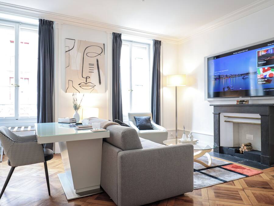 A modern living room with a flat-screen TV, a fireplace, a sofa, a dining table, and a large window with curtains.