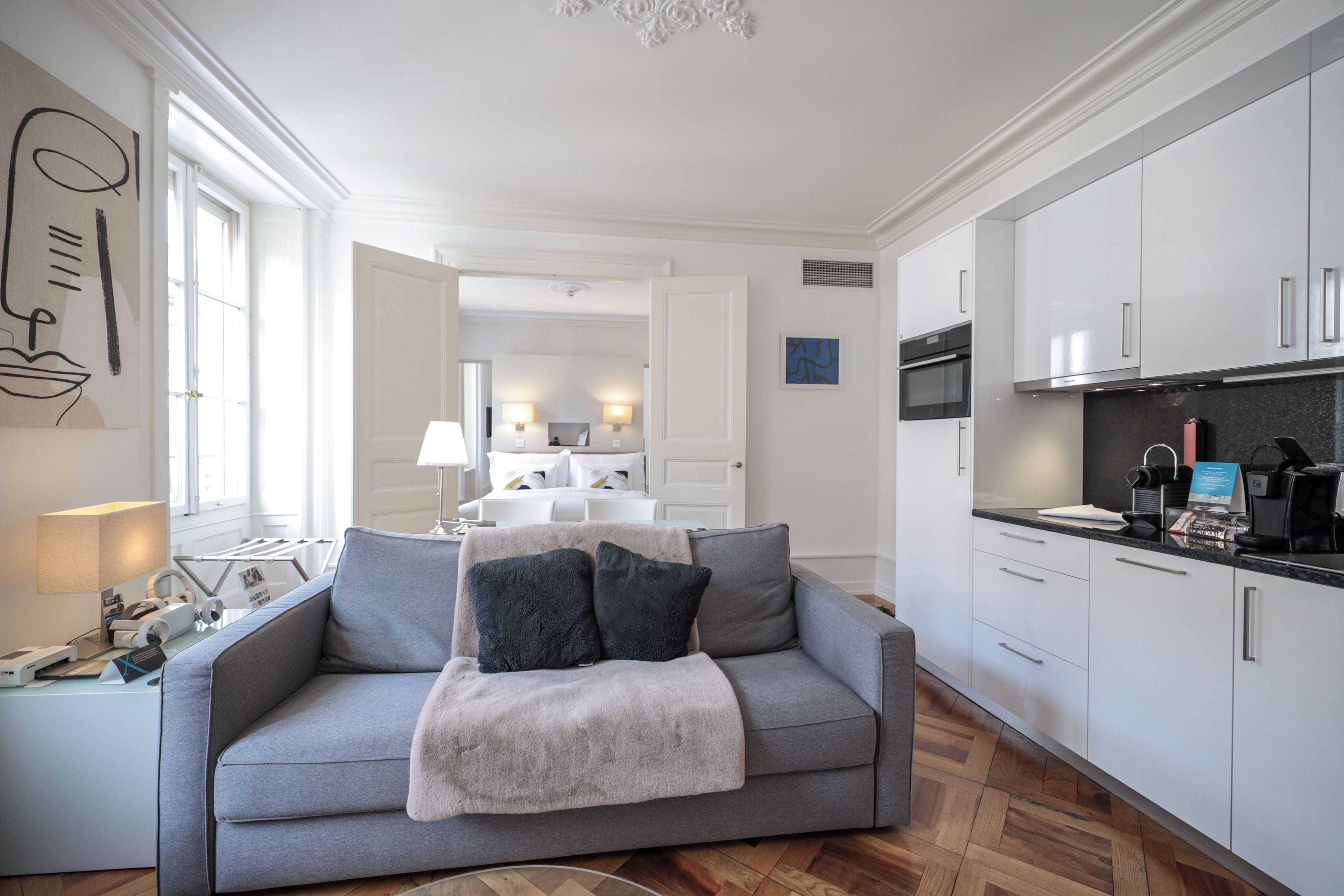 Een moderne woonkamer met een grijze bank, een koffieapparaat en een keuken met witte kasten.