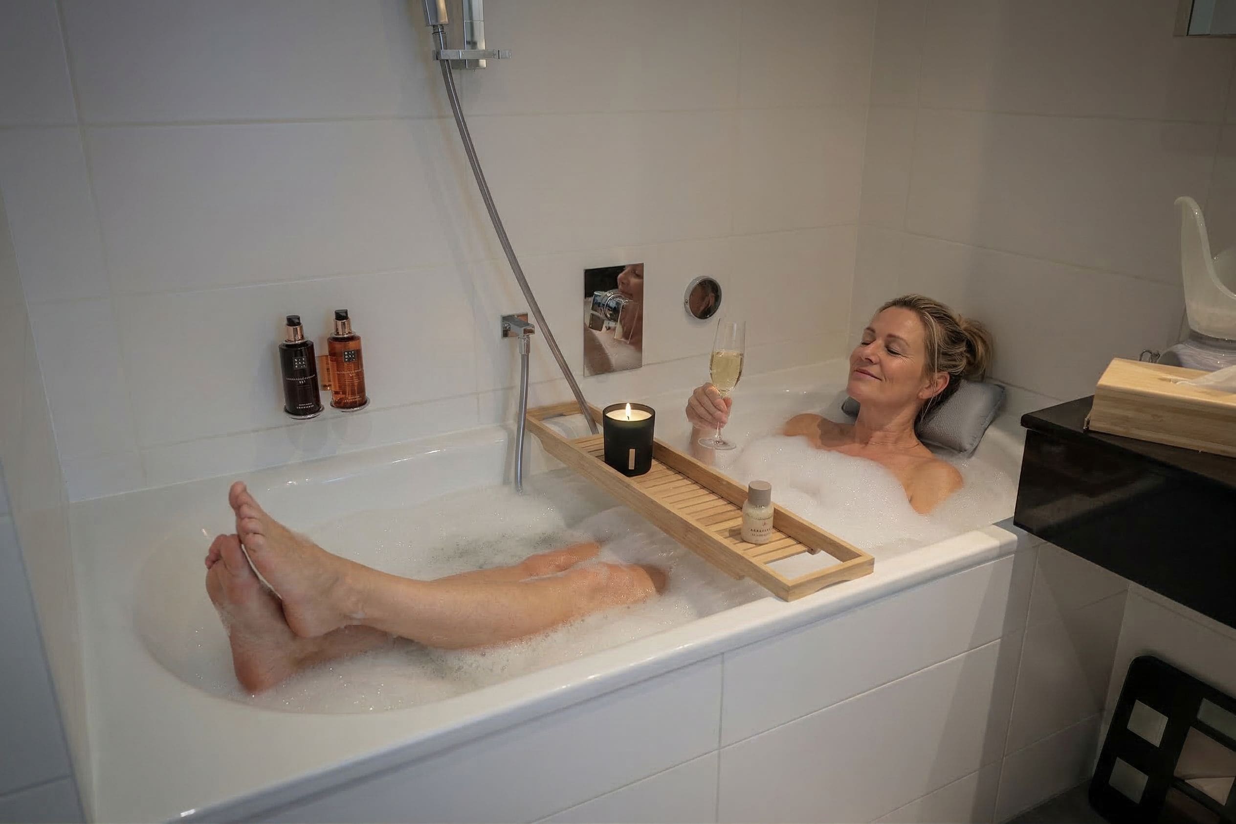 Eine Person entspannt sich in einer Badewanne, auf einem Tablett stehen eine Kerze und ein Glas Wein.