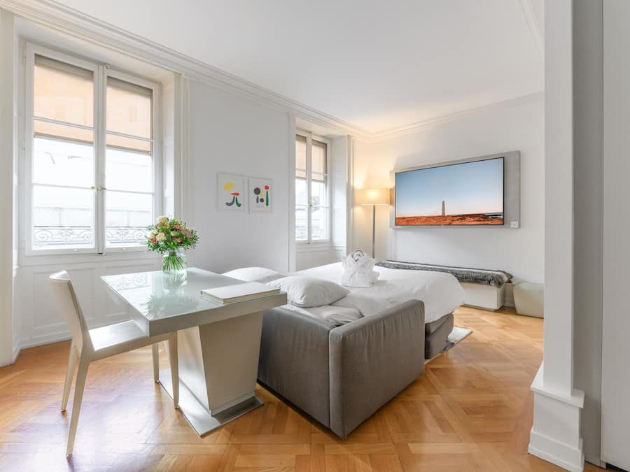 Una habitación de hotel moderna con cama, escritorio y silla. Hay una ventana con vistas a la ciudad, un cuadro en la pared y un jarrón con flores sobre el escritorio.