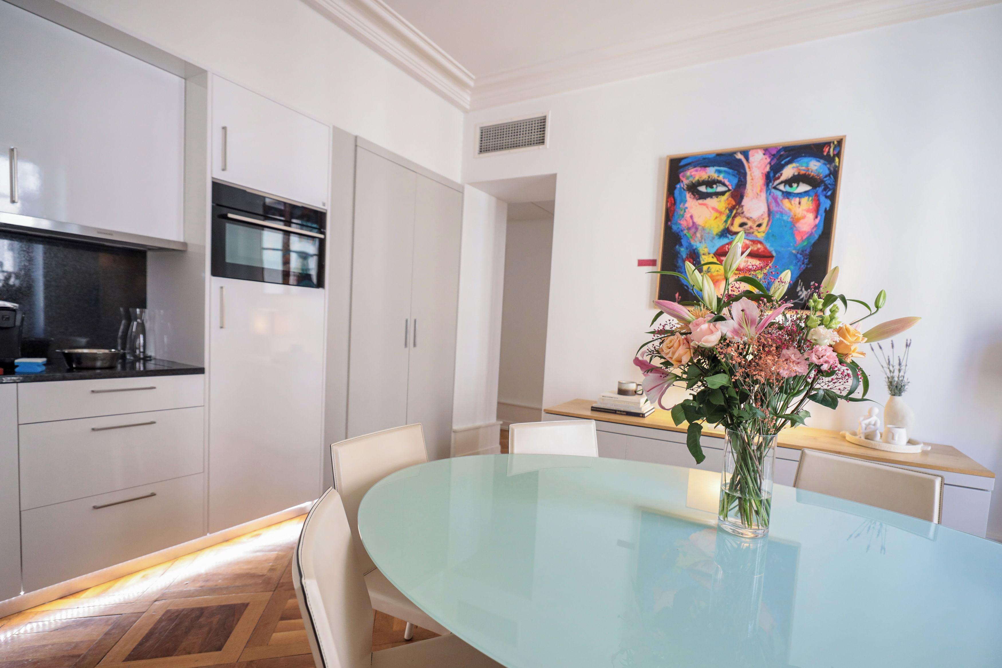 A modern kitchen with white cabinets, a built-in oven, and a dining area with a glass table and chairs.