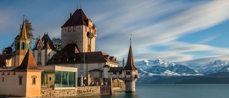 A historic castle with a tower, a glass-walled building, and a view of snow-capped mountains across a calm lake.