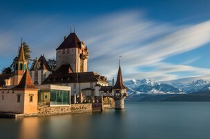 Un castillo histórico con una torre, un edificio de paredes de vidrio y vistas a montañas nevadas sobre un lago tranquilo.