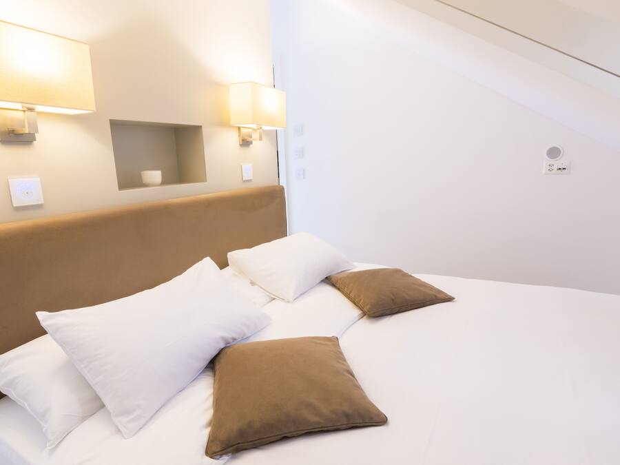 Una cama bien hecha con almohadas blancas y marrones, un cabecero y dos lámparas fijadas a la pared.