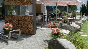 An outdoor seating area with tables and chairs, a fire pit, and a stone wall with stacked logs. - Hotel GARNI Mittenwald (Flums)