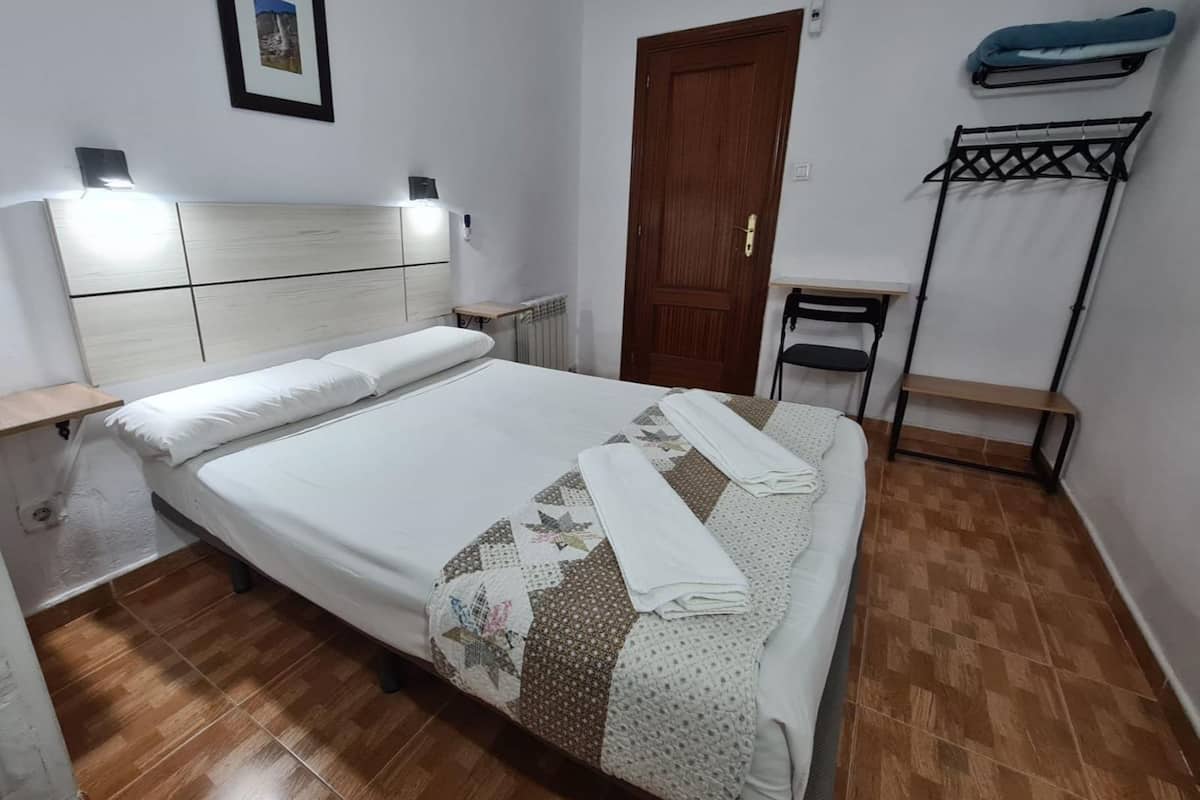 A neatly made bed with a quilt, a wooden headboard, a small bedside table, a radiator, a wooden door, a chair, a bench, and a wall-mounted rack.