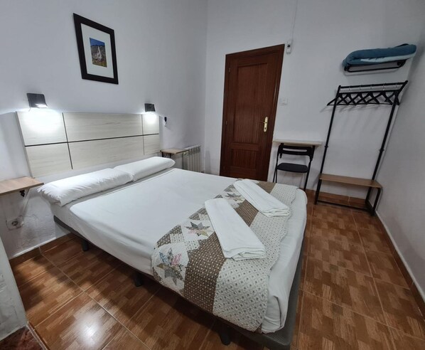 A neatly made bed with a quilt, a wooden headboard, a small bedside table, a radiator, a wooden door, a chair, a bench, and a wall-mounted rack.
