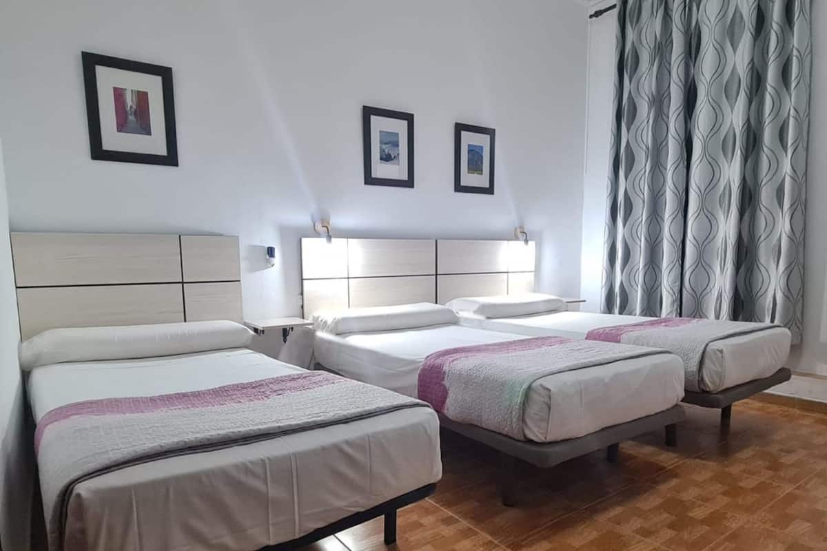 A hotel room with four single beds, a wooden floor, a white headboard, and a patterned curtain.
