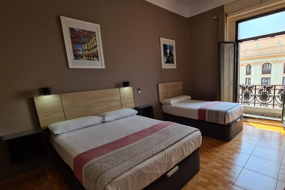 A hotel room with two beds, wooden flooring, and a balcony with a view of buildings.