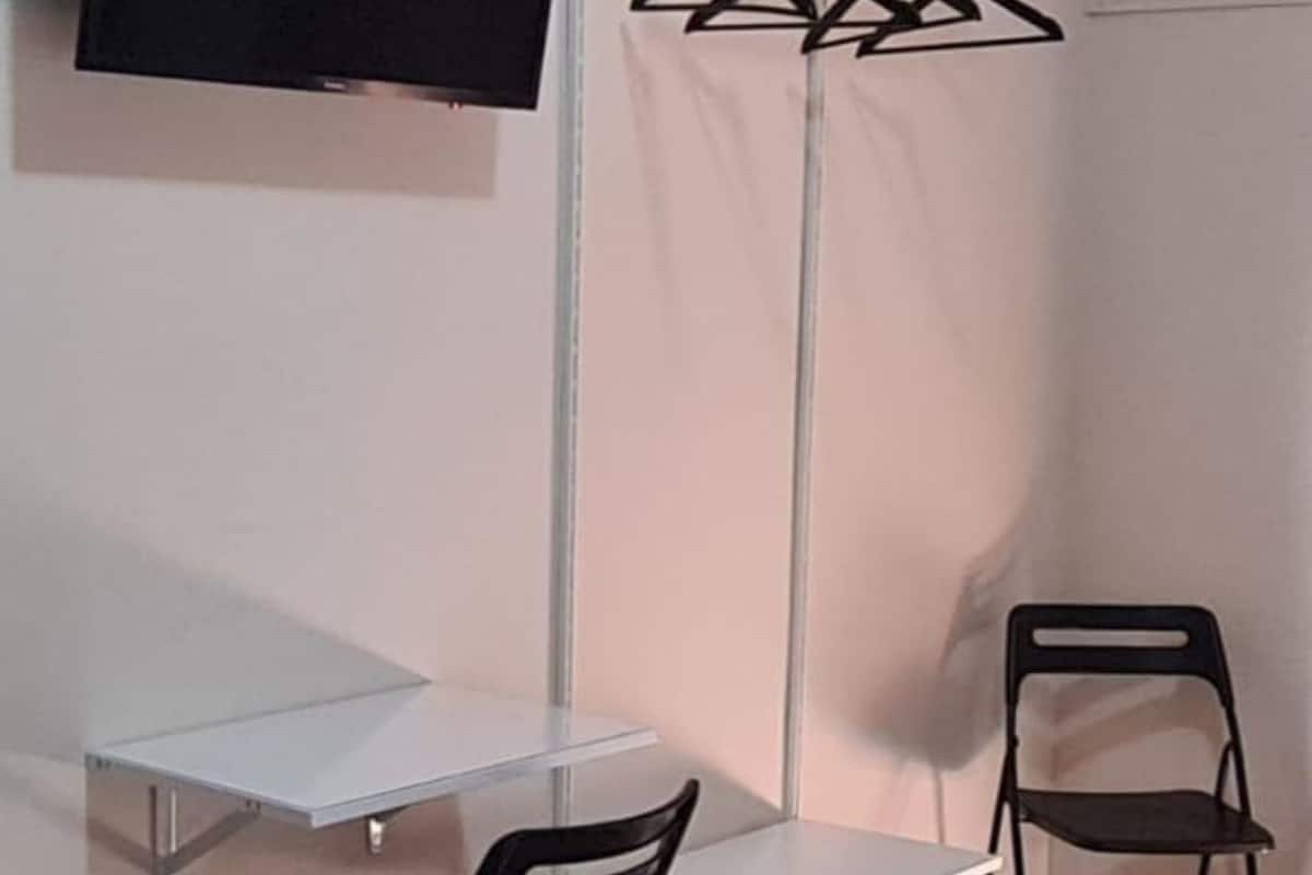 A corner room with a white desk, two black chairs, a TV mounted on the wall, and a clothing rack with hangers.