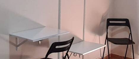 Een hoekappartement met een wit bureau, twee zwarte stoelen, een aan de muur bevestigde televisie en een kledingrek met hangers.