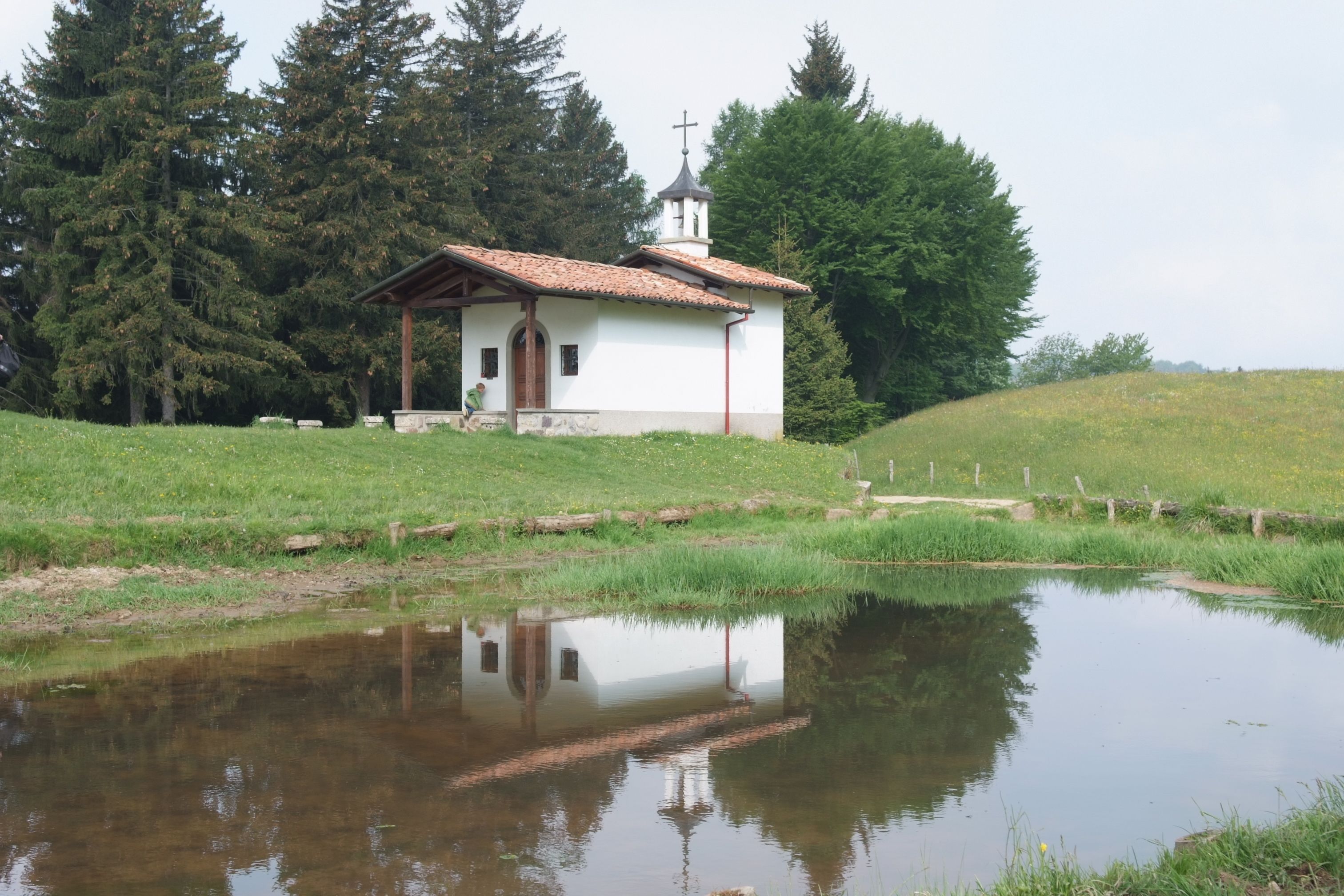 A small white church with a tiled roof, surrounded by a grassy field and a body of water reflecting its structure.