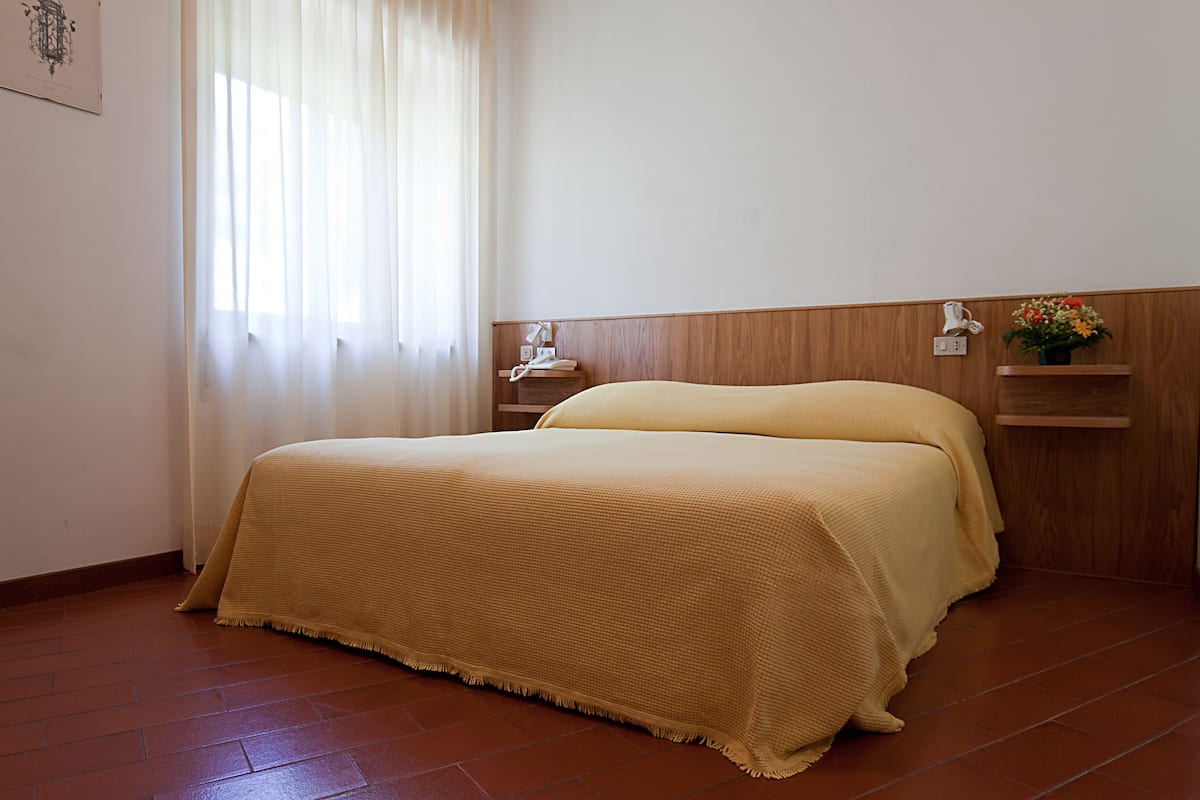 A hotel room with a bed, a nightstand, a wall-mounted shelf, and a window with curtains.