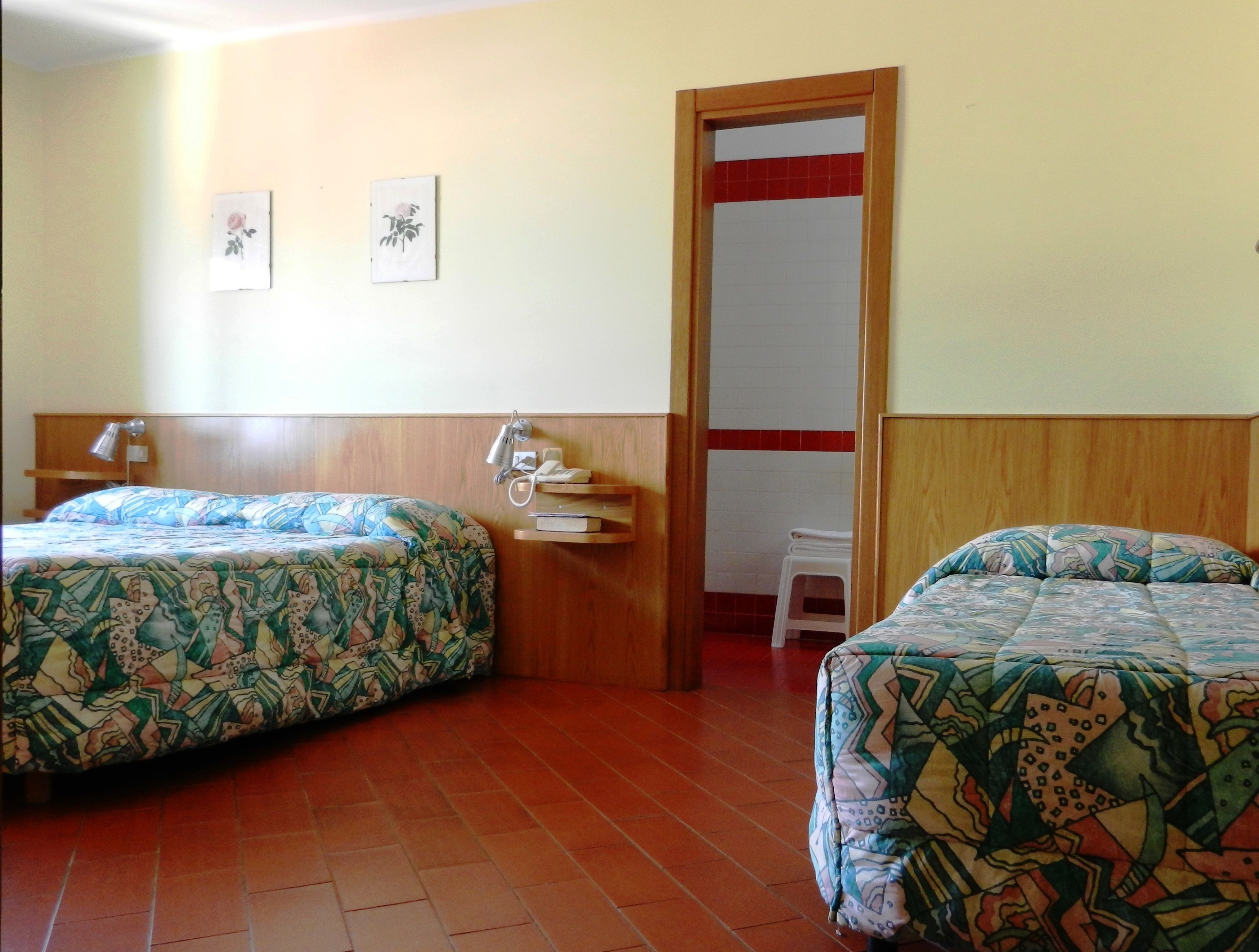 A hotel room with two beds, a wooden headboard, a small table with a lamp, and a red and white tiled wall.