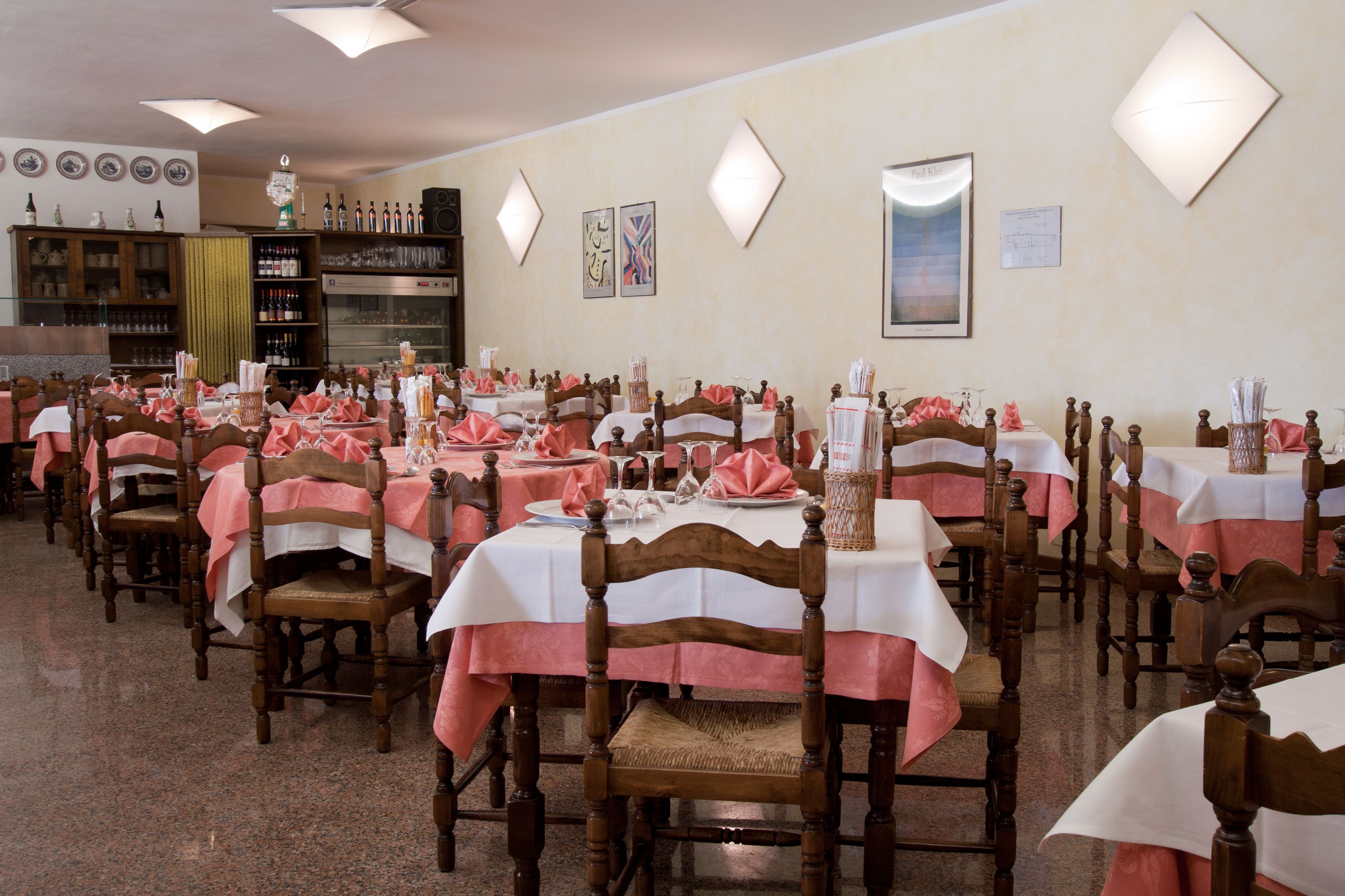 Un restaurante con varias mesas cubiertas con manteles blancos y rosas, sillas de madera y lámparas fijadas a la pared.
