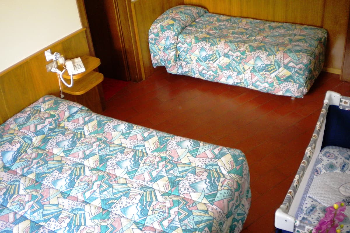 A hotel room with two beds, a wooden headboard, a small wooden table with a telephone, and a wall-mounted light switch.