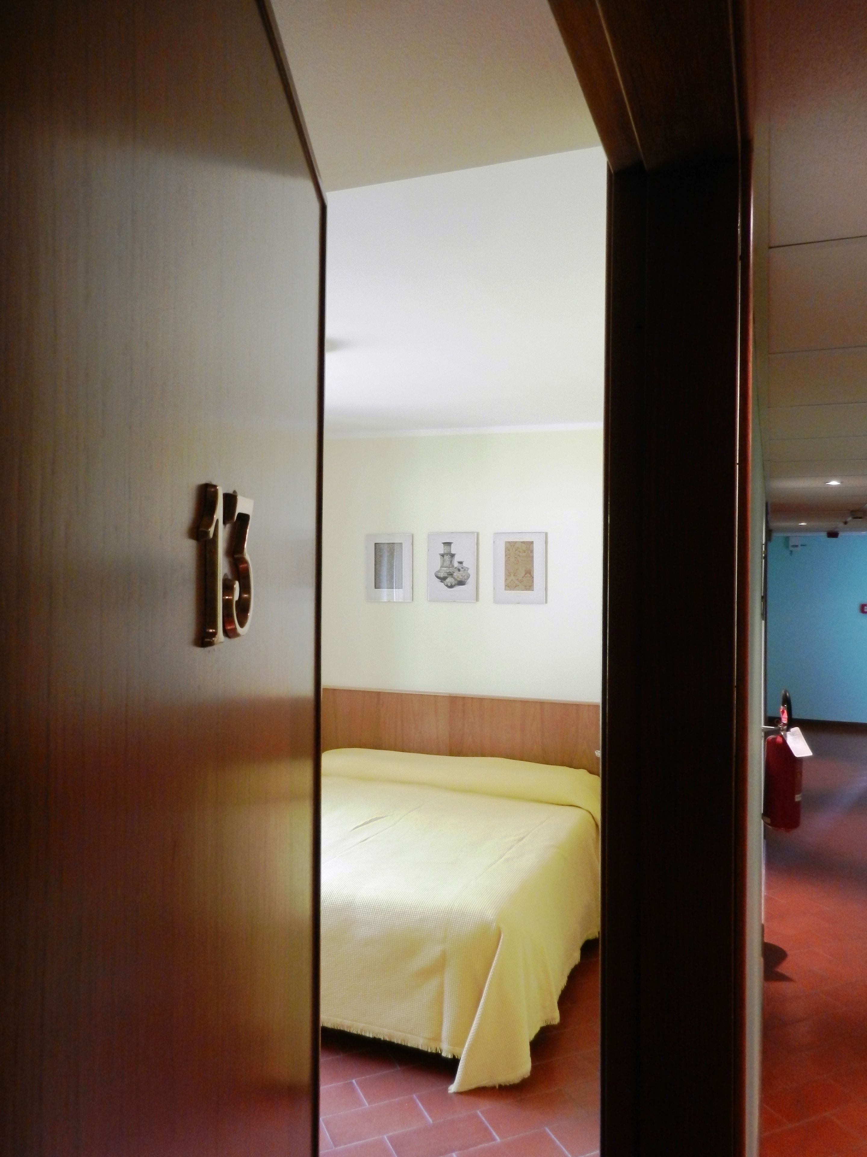 Una habitación de hotel con una cama, una puerta con un número y tres cuadros en la pared.