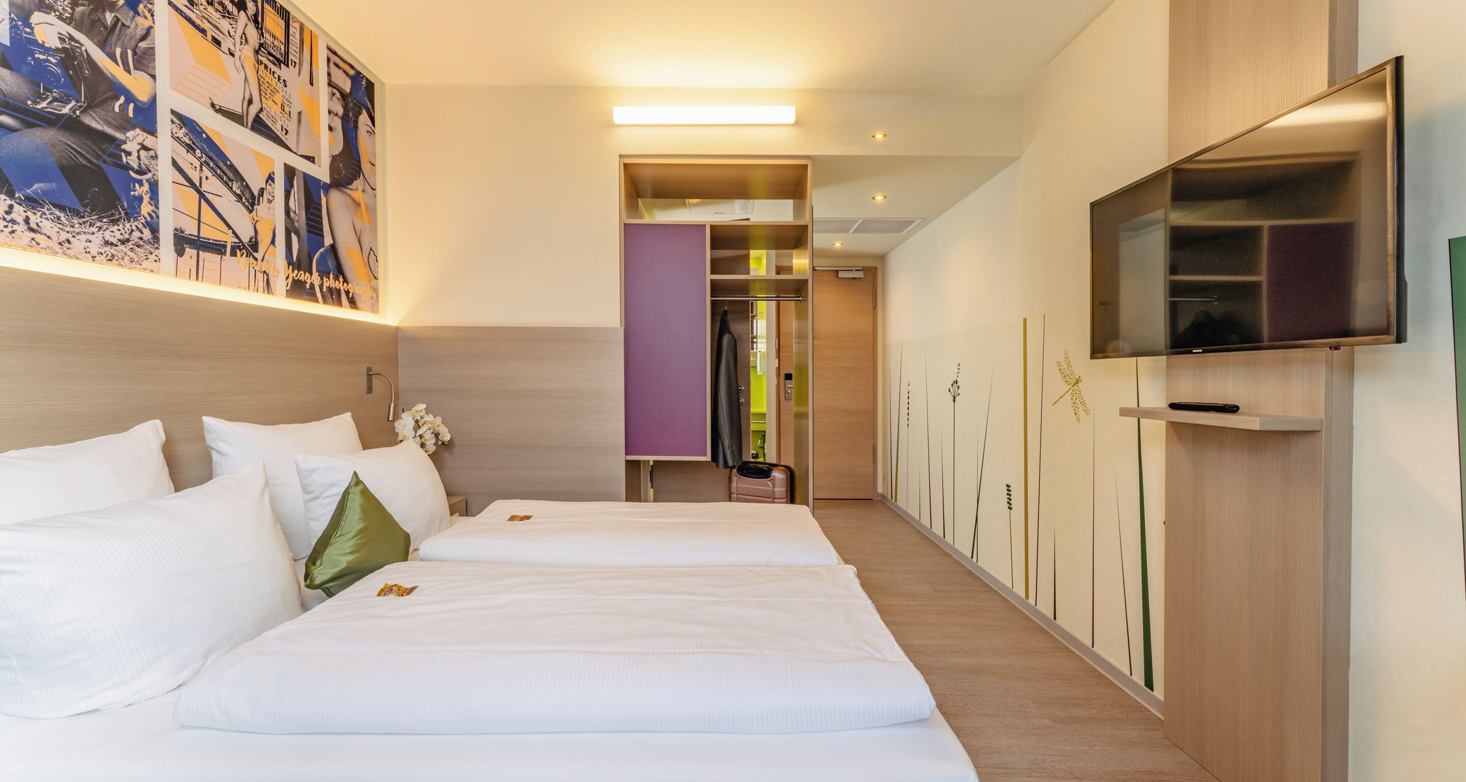 A hotel room with two beds, a TV, a hallway with a mirror, and a wall with a floral design.