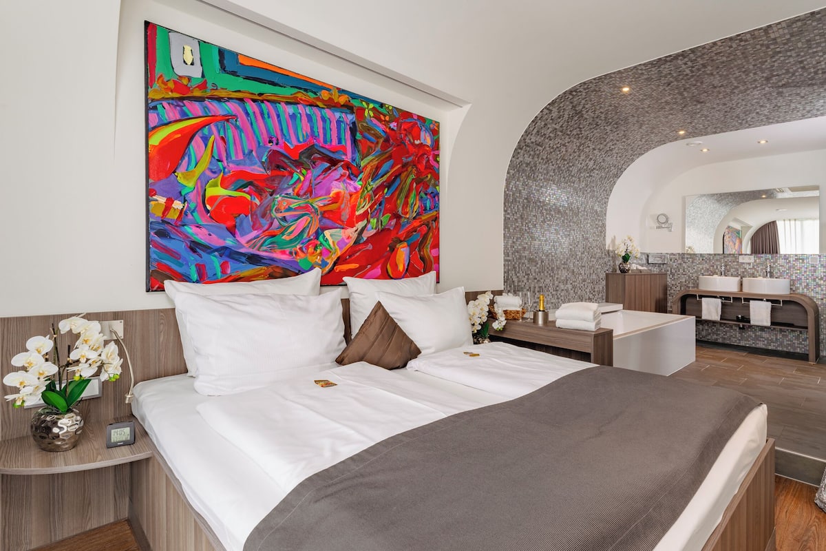A modern hotel room with a large bed, a bathroom with a mirror, and a colorful abstract painting on the wall.