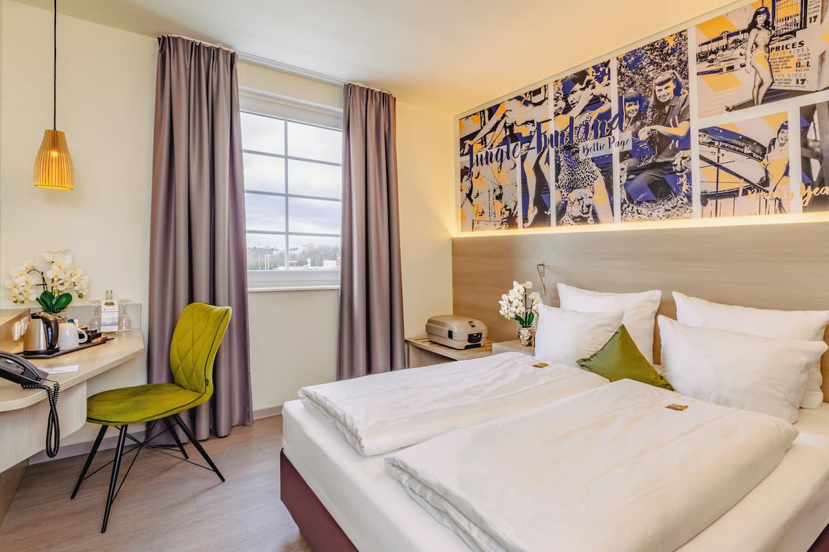 A hotel room with a bed, a desk with a phone, a chair, a nightstand with a lamp, and a wall with a poster.