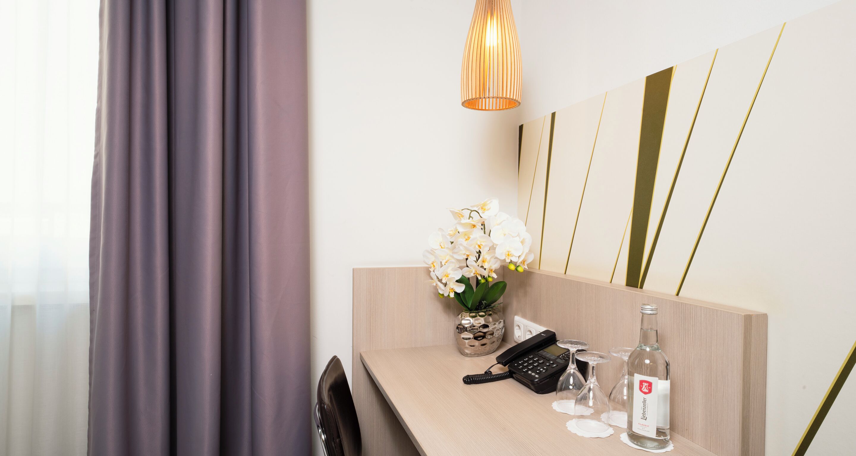 Habitación de hotel con un escritorio de madera, un teléfono, una botella de agua y un florero con flores blancas.