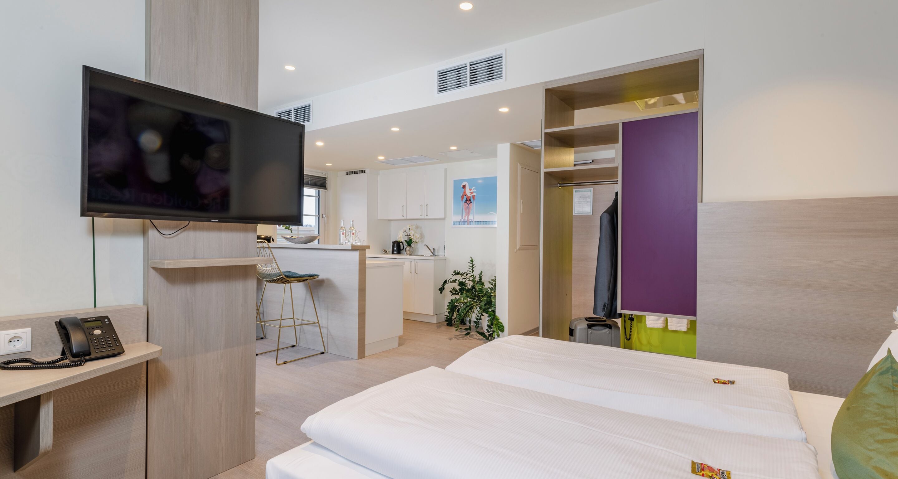 A hotel room with a bed, a desk with a phone, a TV, a kitchenette, and a bathroom.