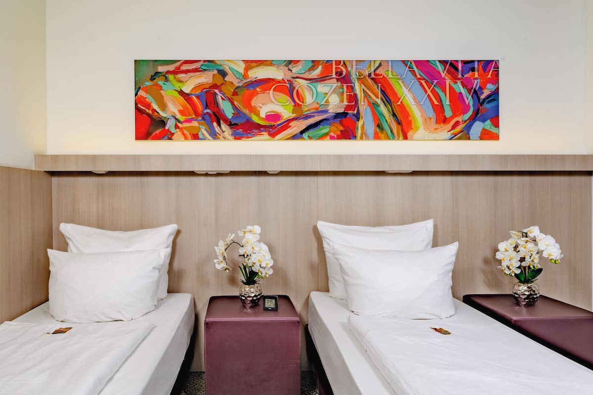 A hotel room with two beds, white bedding, and a colorful abstract painting above.