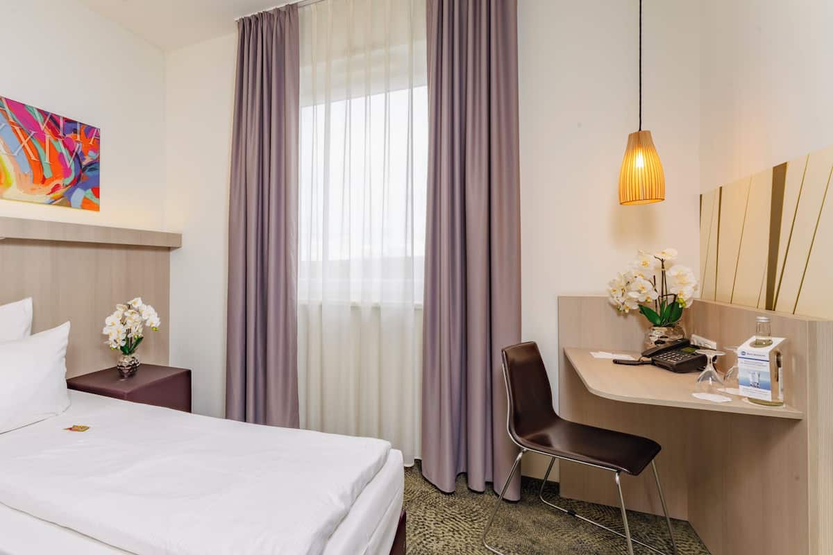 A hotel room with a bed, a desk with a chair, a vase of flowers, a painting on the wall, and a window with curtains.