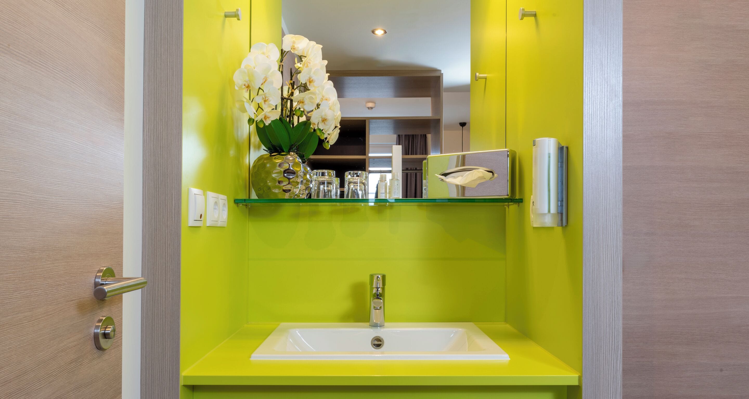 Ein Badezimmer mit einem gelben Waschbecken, einer Glasablage für Toilettenartikel und einer Vase mit weißen Blumen.