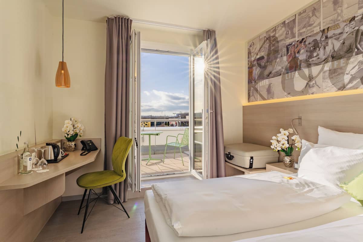 A hotel room with a bed, a desk with a chair, a TV, a window with curtains, and a balcony with chairs.