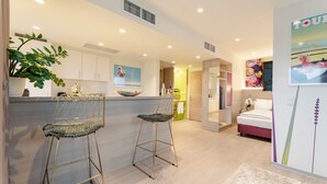 A modern hotel room with a kitchen area, two bar stools, a bed, and a large poster on the wall.