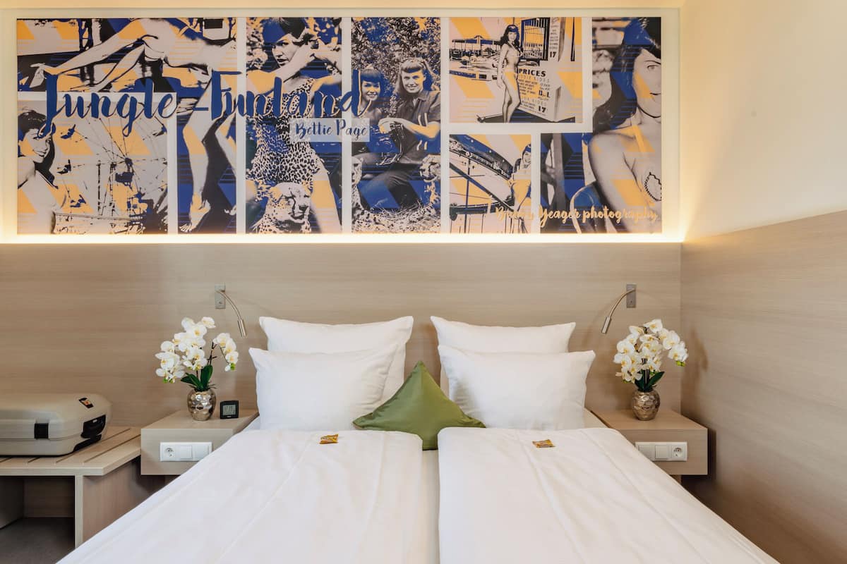 A hotel room with a bed, two bedside tables, a wall art piece, and a small desk with a printer.