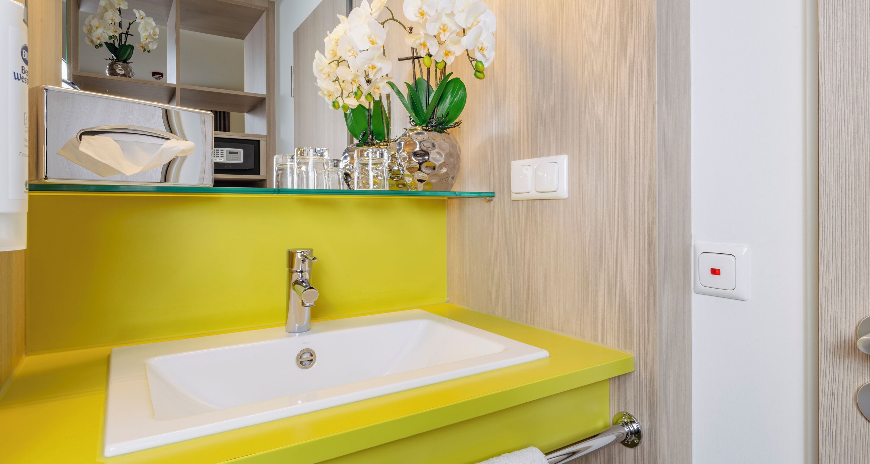 A bathroom with a yellow sink, white faucet, and a glass vase with white flowers on the shelf above.
