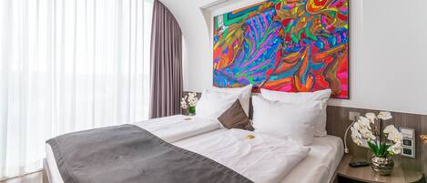 A hotel room with a large bed, white linens, a colorful abstract painting on the wall, a vase of white flowers, and a nightstand with a remote control.