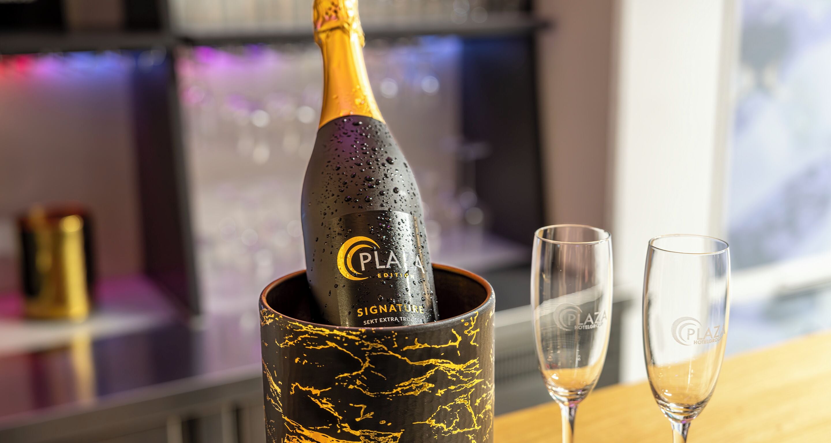 A champagne bottle in a black and gold mug with two glasses beside it.