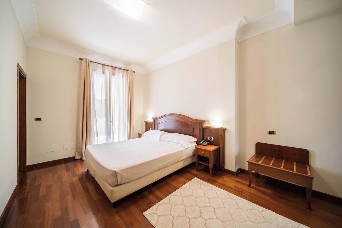 Un dormitorio con cama, mesita de noche, lámpara y un banco.