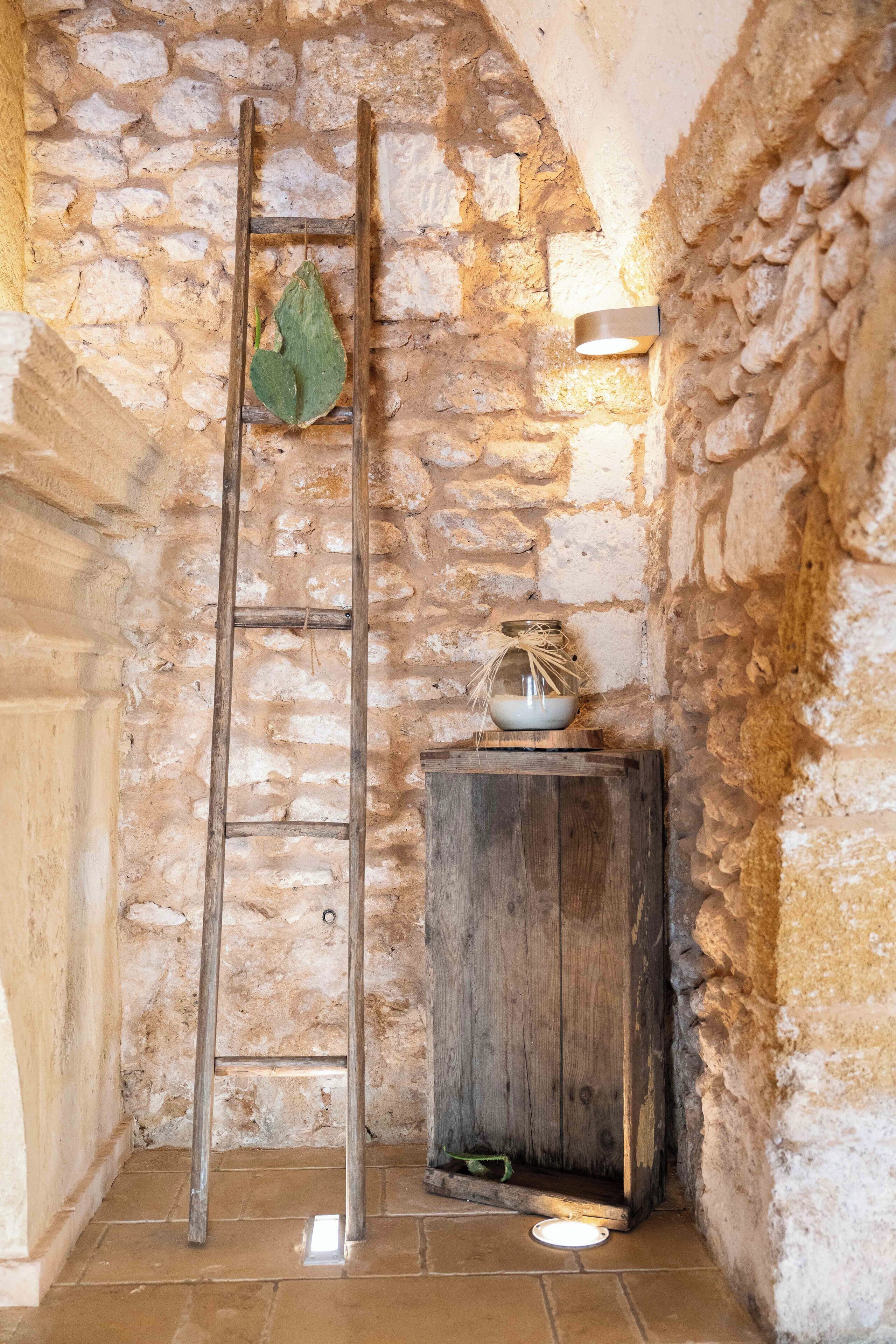 A stone-walled room with a wooden ladder leaning against the wall, a small wooden cabinet with a jar on top, and a light fixture on the wall.