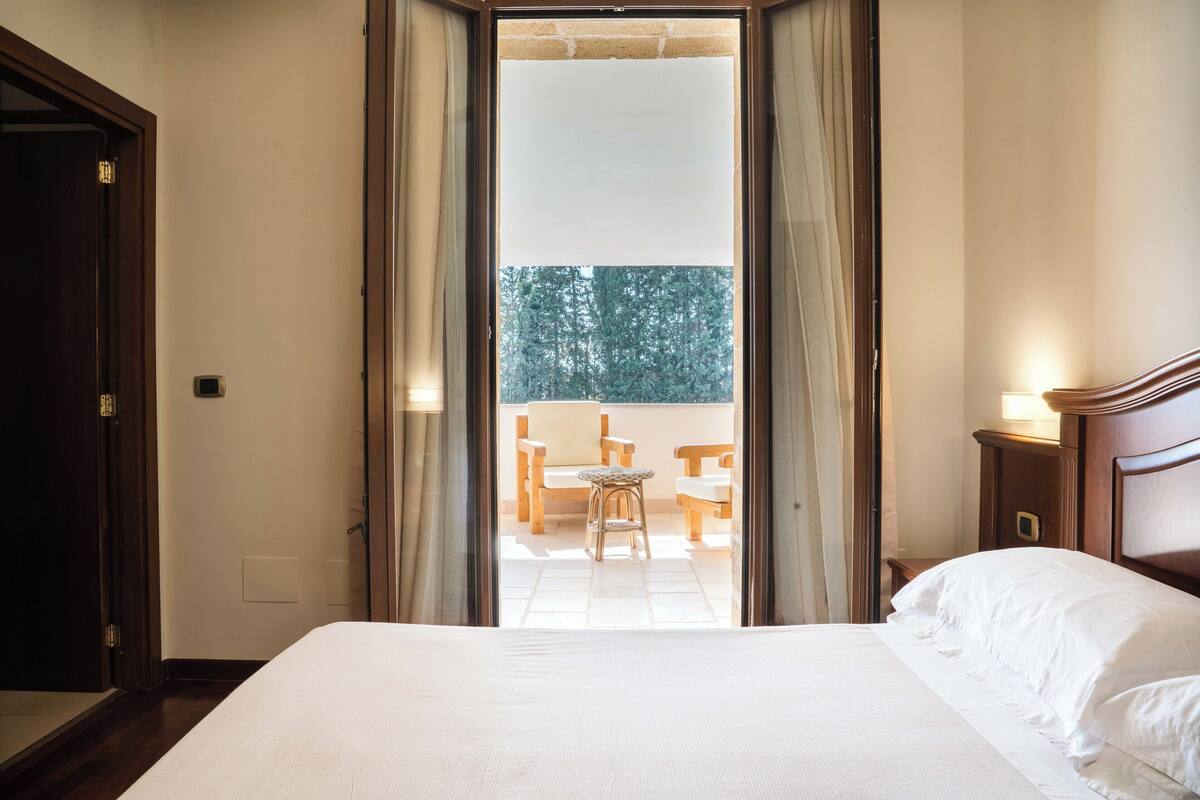 Habitación de hotel con cama, mesita de noche de madera, ventana con cortinas y vista a un balcón con sillas y una mesa.
