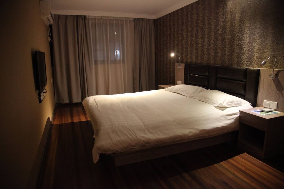 A hotel room with a large bed, a bedside table with a lamp, a wall-mounted TV, and a window with curtains.