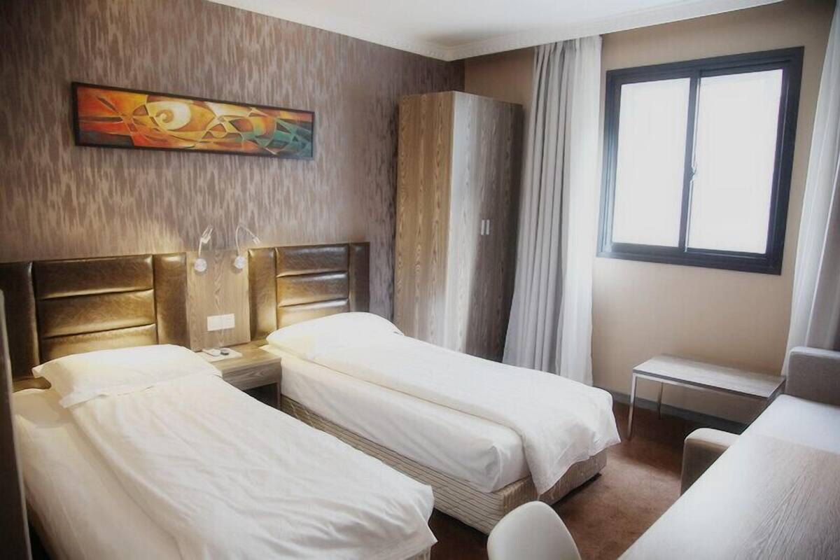 A hotel room with two single beds, a wooden wardrobe, a small table, and a window with curtains.
