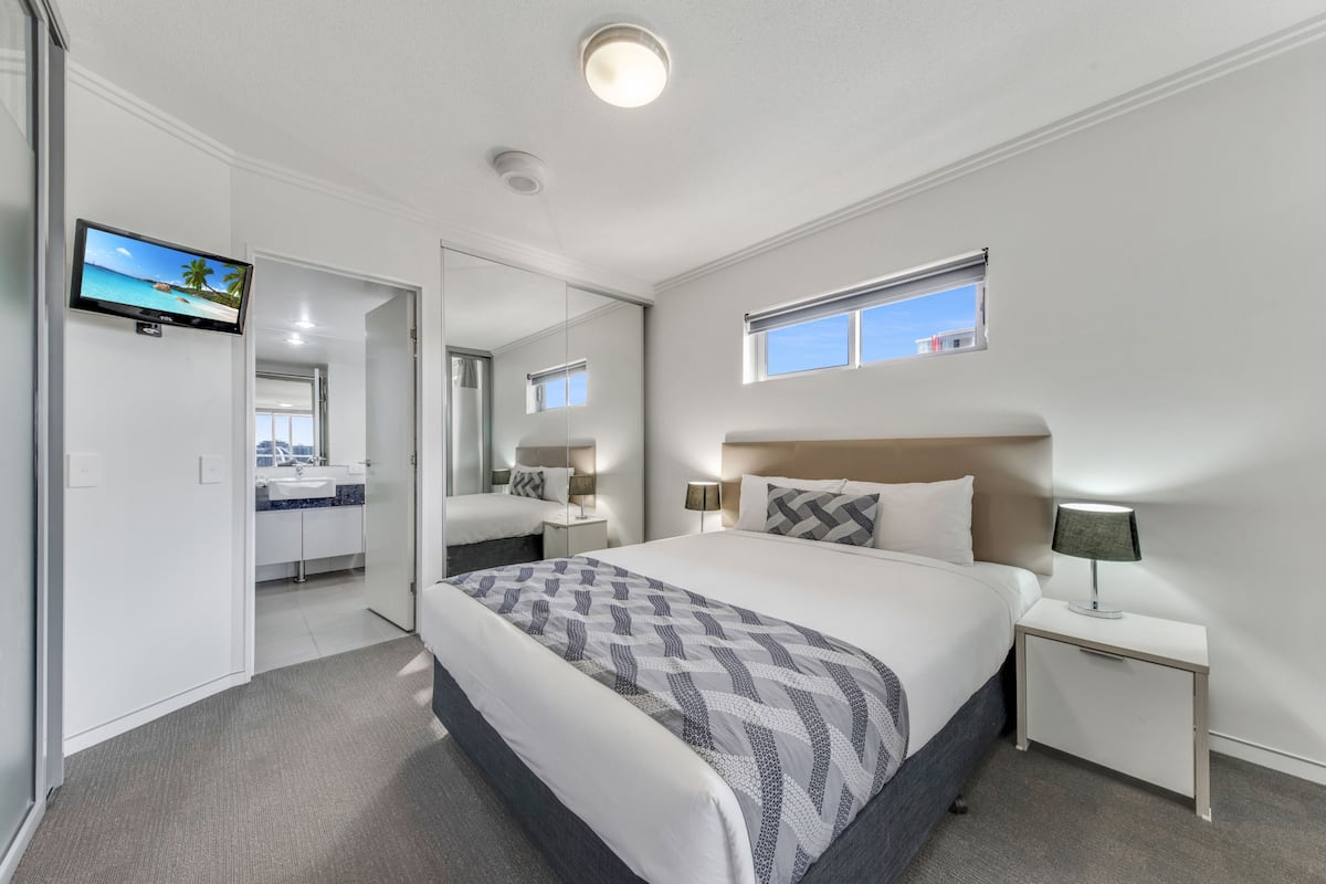 A modern hotel room with a large bed, a TV mounted on the wall, a small window, and a bathroom visible through an open door.