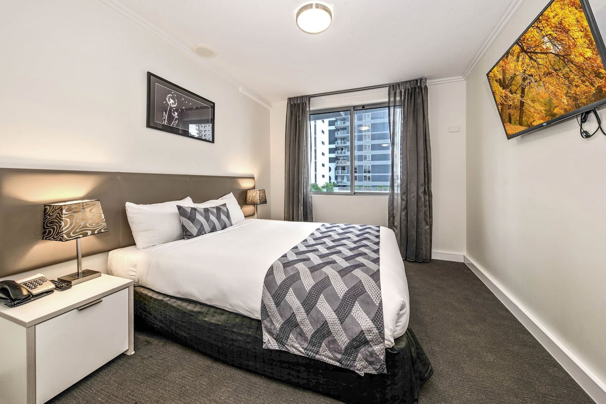 A hotel room with a bed, nightstand, lamp, and a large window with curtains.