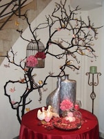 Een tafel met een rode tafelkleed, een decoratieve boom met takken, een vogelkooi en een vaas met bloemen.