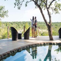 Una pareja de pie en una terraza de madera con vista a una piscina y un paisaje exuberante.