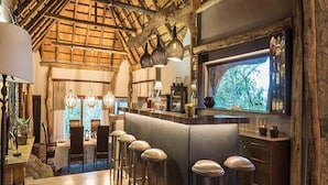 A bar area with a high ceiling and exposed wooden beams, a bar counter with stools, and a view of trees through a window. - Leopard Hills (Bushbuckridge)