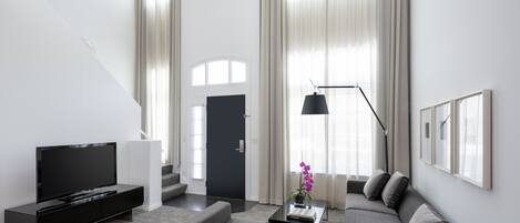 Ett modernt vardagsrum med en platt-TV, en soffa, ett soffbord med en bok och en vas med blommor samt en trappa som leder till ett annat rum.