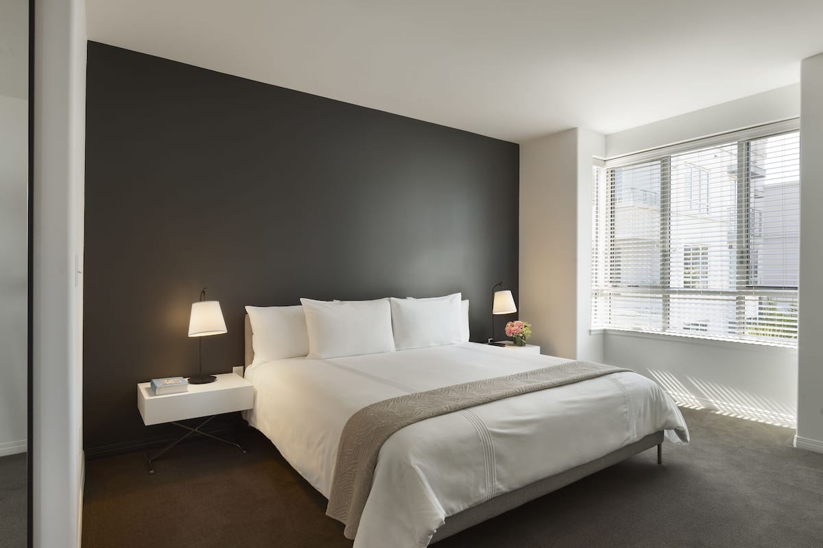 A modern hotel room with a large bed, two bedside tables, a nightstand, a window with blinds, and a view of the cityscape.