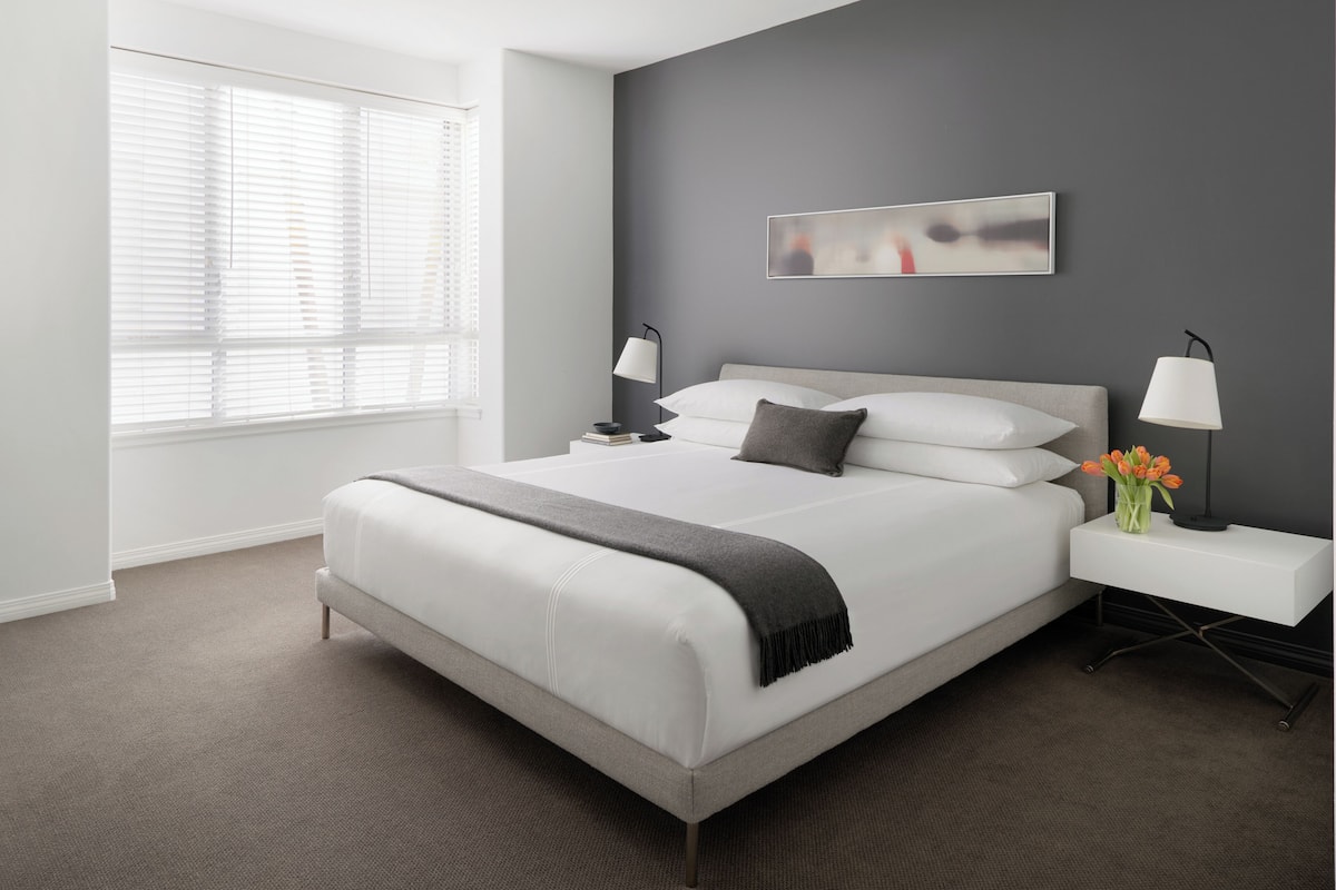 A modern bedroom with a large bed, bedside tables, a nightstand, a window with blinds, and a wall-mounted abstract art piece.